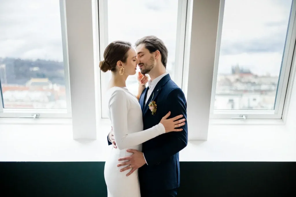 A bride and groom stand closely together, embracing and touching foreheads, in front of large windows with a cityscape view—the perfect moment for weddings and cherished social gatherings.